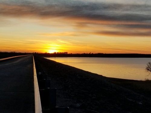 A serene sunset over a calm lake, with a road leading alongside the water and colorful clouds in the sky.