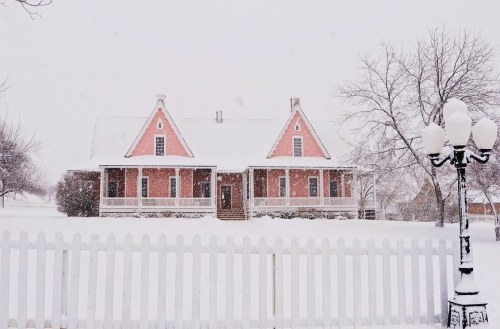 A pink house covered in snow, surrounded by a white picket fence, with a lamppost in the foreground on a snowy day.