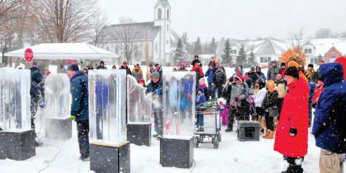 A crowd watches as ice sculptures are created during a snowy outdoor event, with a church in the background.