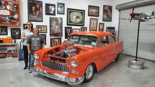 Two women stand beside a bright orange classic car with an exposed engine, surrounded by framed memorabilia in a garage.