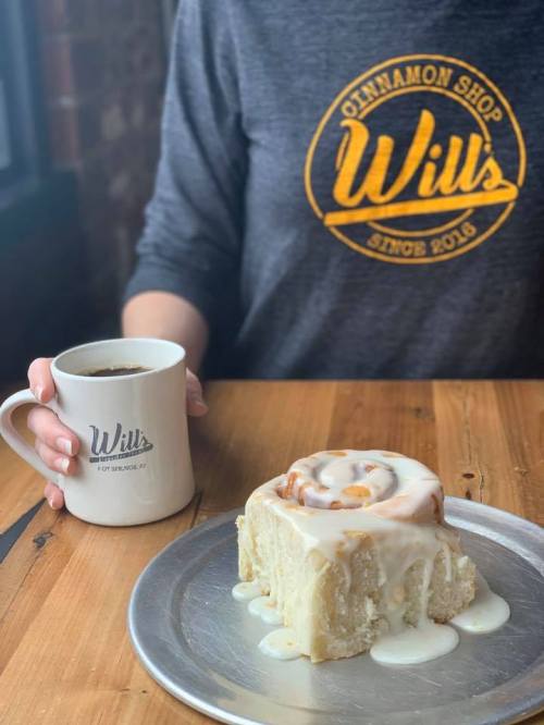 A person holds a coffee mug next to a cinnamon roll on a plate, with a wooden table and a shop logo in the background.