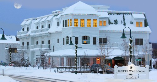 A snowy scene featuring Hotel Walloon, a charming white building adorned with holiday lights and a full moon in the sky.