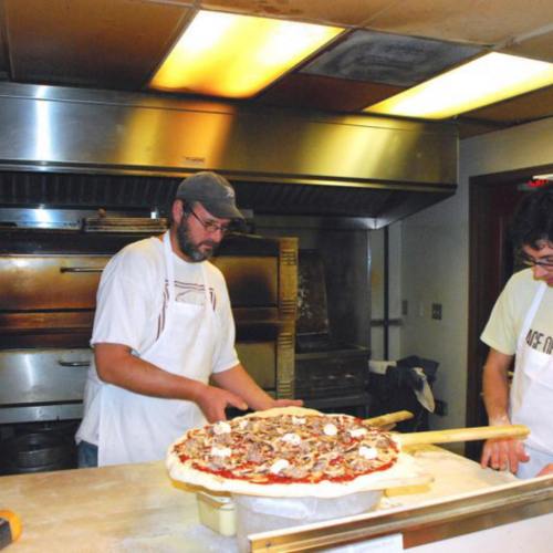A chef prepares a large pizza in a kitchen, with another person nearby and an oven in the background.
