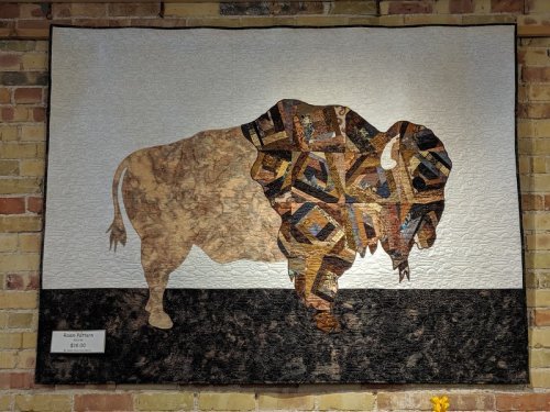 A quilted artwork of a bison, featuring intricate patterns and textures, displayed on a brick wall.