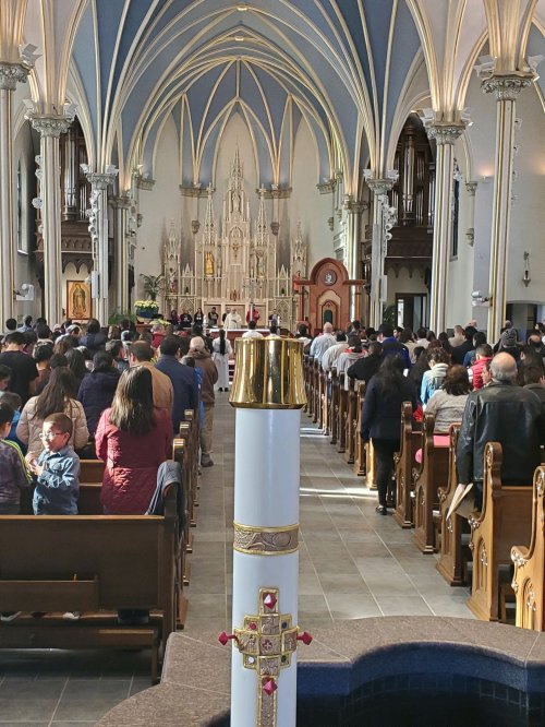 A view of a church service with a congregation gathered, featuring ornate architecture and a prominent candle in the foreground.