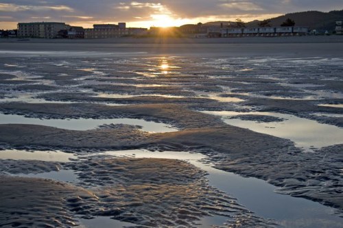 Sunset over a beach with rippled sand and reflections, buildings in the background, creating a serene coastal scene.