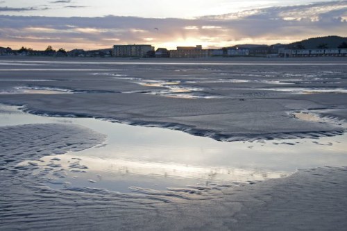 A serene beach at sunset, with reflections in shallow water and distant buildings silhouetted against the sky.