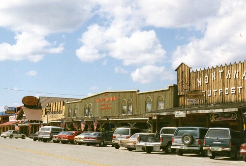 A row of rustic storefronts with wooden facades and parked cars under a blue sky with fluffy clouds.