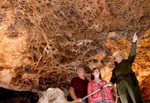 A park ranger points out cave formations to two visitors, who are observing the intricate ceiling of the cave.