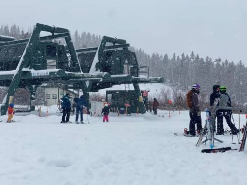 Skiers and snowboarders gather near a ski lift in a snowy mountain landscape. Snow is falling heavily.
