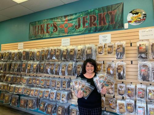 A woman stands in a store holding packages of jerky, with a wall of jerky products displayed behind her.