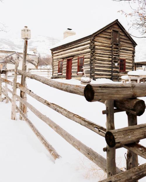 A rustic log cabin surrounded by a wooden fence, blanketed in snow, with mountains in the background.