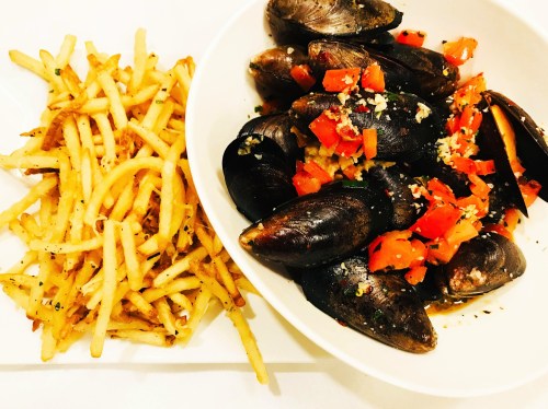 A bowl of mussels topped with diced tomatoes and garlic, served alongside a plate of crispy French fries.