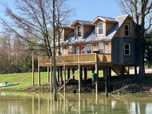 A wooden house on stilts by a calm lake, surrounded by trees and greenery, with a kayak nearby.