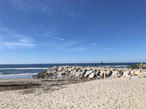 A sandy beach with a rocky jetty, people walking along the shore, and clear blue skies above.