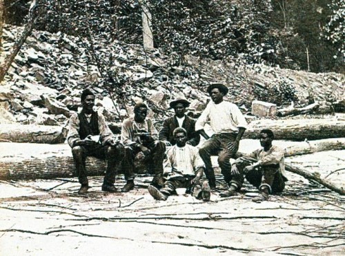 A historic black-and-white photo of seven men sitting on logs in a forested area, surrounded by rocks and trees.