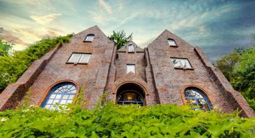 A unique brick building with a triangular roof, surrounded by greenery and under a dramatic sky.