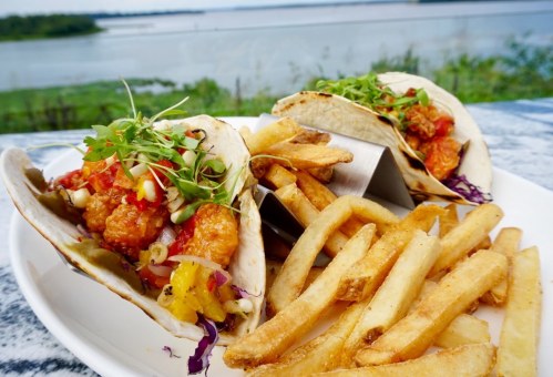 Two tacos filled with colorful toppings, served with a side of golden fries, against a scenic waterfront backdrop.
