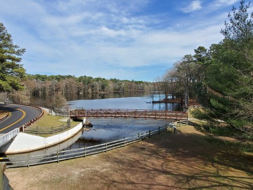 A serene lake view with a wooden bridge, surrounded by trees and a winding road under a clear blue sky.