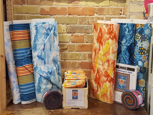 Colorful fabric rolls in various patterns, stacked beside a display of quilting supplies and a rainbow of fabric strips.