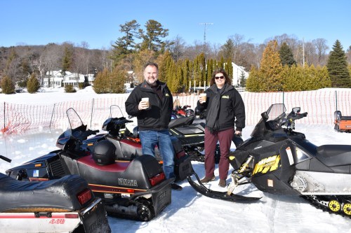 Two people stand beside snowmobiles on a snowy landscape, holding drinks and smiling in the winter sun.