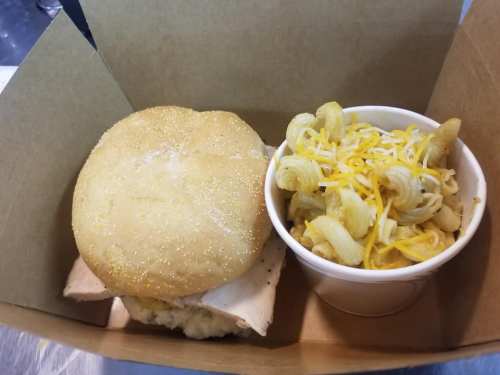 A takeout box containing a sandwich with a golden bun and a side of macaroni and cheese topped with shredded cheese.