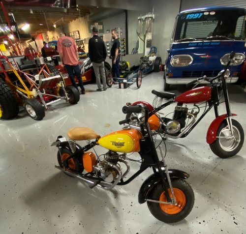Two vintage mini bikes in a garage with people and various vehicles in the background.