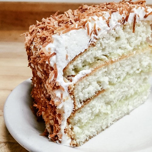 A slice of coconut cake topped with toasted coconut flakes, served on a small plate.