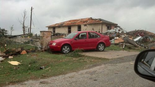 A red car parked near a damaged house with a collapsed roof and debris scattered around, under a cloudy sky.
