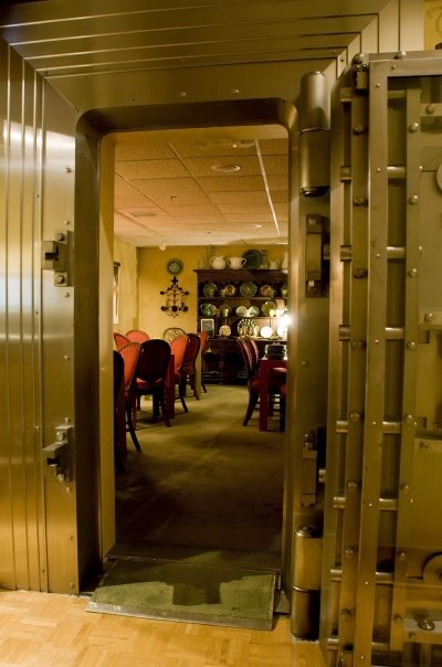 A large, metallic vault door opens to reveal a dining room with a table and decorative plates on the wall.