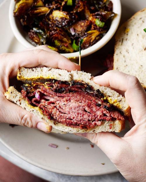 A person holds a thick pastrami sandwich with mustard, alongside a bowl of crispy Brussels sprouts.