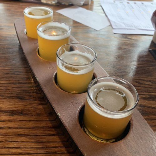 A wooden flight board with four small glasses of light-colored beer on a rustic table.