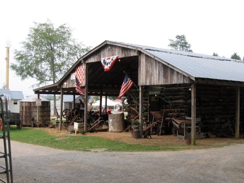 A rustic barn with American flags, showcasing vintage farming equipment and a gravel path in a rural setting.