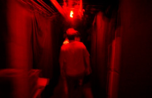 A dimly lit hallway with red lighting, featuring blurred figures walking through a dark space.