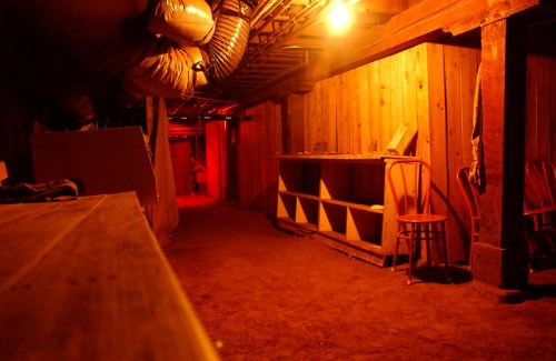 Dimly lit basement corridor with wooden walls, shelves, and chairs, illuminated by a warm orange glow.