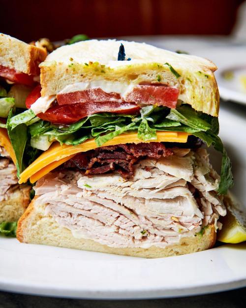 A close-up of a layered sandwich with turkey, bacon, cheese, lettuce, and tomato between two slices of bread.