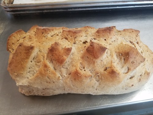 A freshly baked loaf of bread with a golden-brown crust and a textured surface, resting on a metal tray.