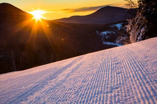 A snowy landscape at sunset, with ski tracks leading down a slope and mountains in the background.