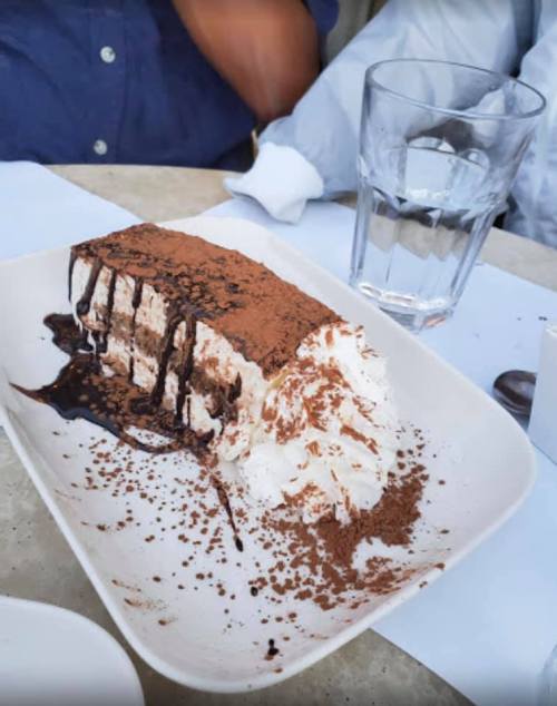 A slice of chocolate dessert topped with whipped cream and cocoa powder, served on a white plate with a glass of water nearby.