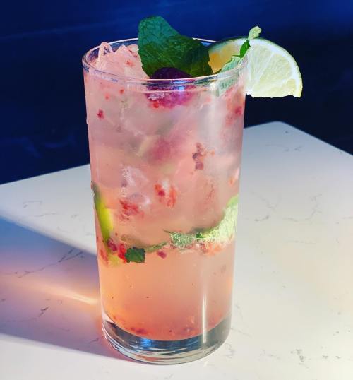 A refreshing cocktail with mint, lime, and berries, served over ice in a tall glass on a marble surface.