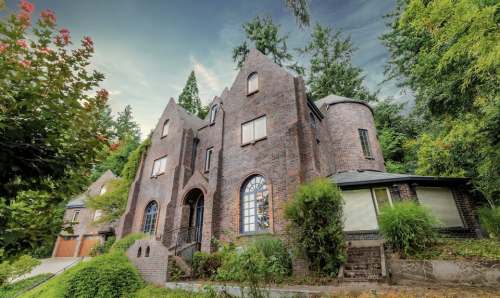 A unique brick house with gothic architecture, surrounded by lush greenery and tall trees under a cloudy sky.
