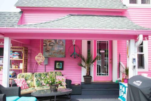 A vibrant pink house with a cozy porch, featuring colorful decor, plants, and a peace sign hanging by the entrance.