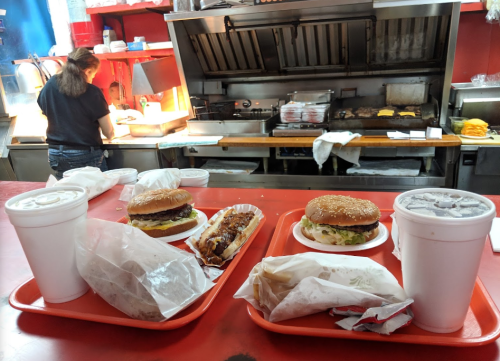 A counter with two trays of burgers and drinks, with a cook preparing food in the background.