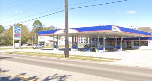 A gas station with a blue and red canopy, featuring fuel pumps and a convenience store, located on a busy street.