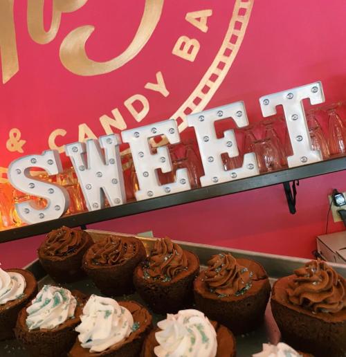A display of chocolate cupcakes with frosting in front of a bright pink wall featuring the word "SWEET" in marquee letters.