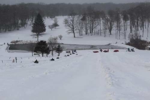 A snowy hill with sledders in the distance, surrounded by trees and a frozen pond. Overcast sky.