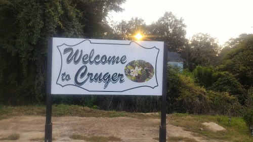 A sign reading "Welcome to Cruger" with a floral design, set against a backdrop of trees and a sunset.