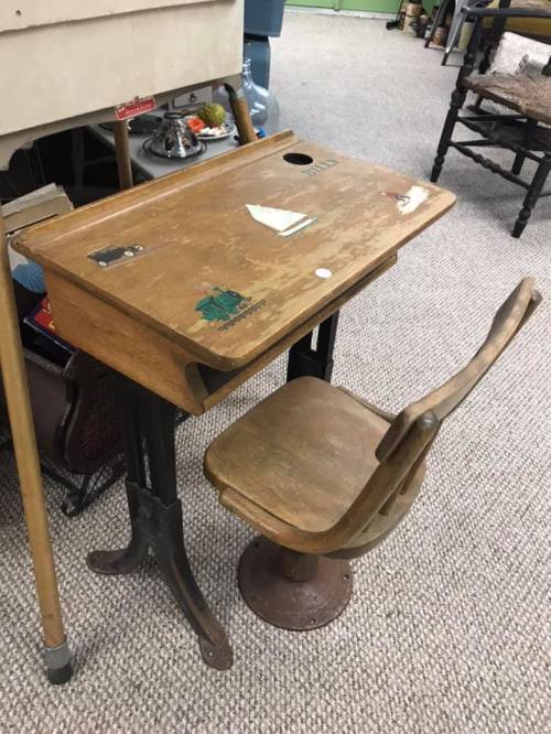 A vintage wooden school desk with a chair, featuring stickers and a hole for an ink well, set on a carpeted floor.