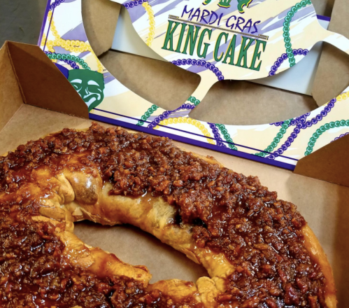 A colorful Mardi Gras king cake with a shiny topping, placed in a box with a decorative mask design.