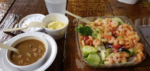 A plate with a shrimp salad, a cup of beans, and a small serving of cornbread on a wooden table.